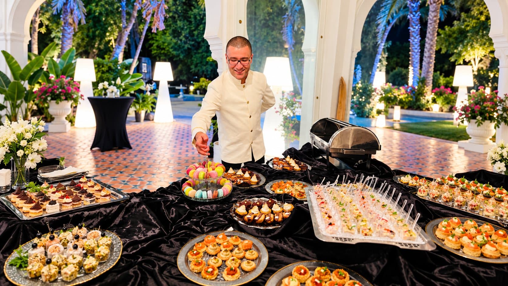 Un chef dispose des mignardises sur un buffet cocktail drapé de noir, en soirée sous des arcades.