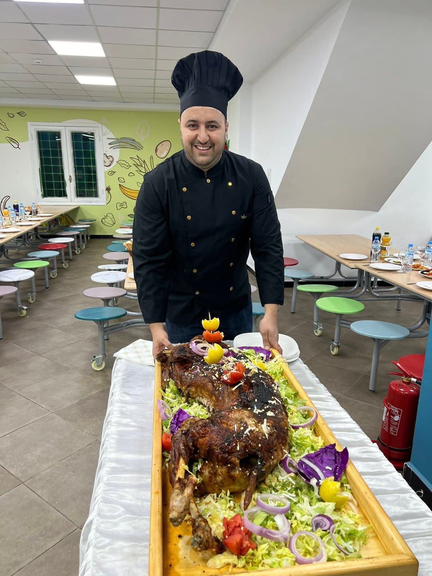 Un chef en veste noire devant un méchoui dressé sur un plateau garni de salade et d'oignons.