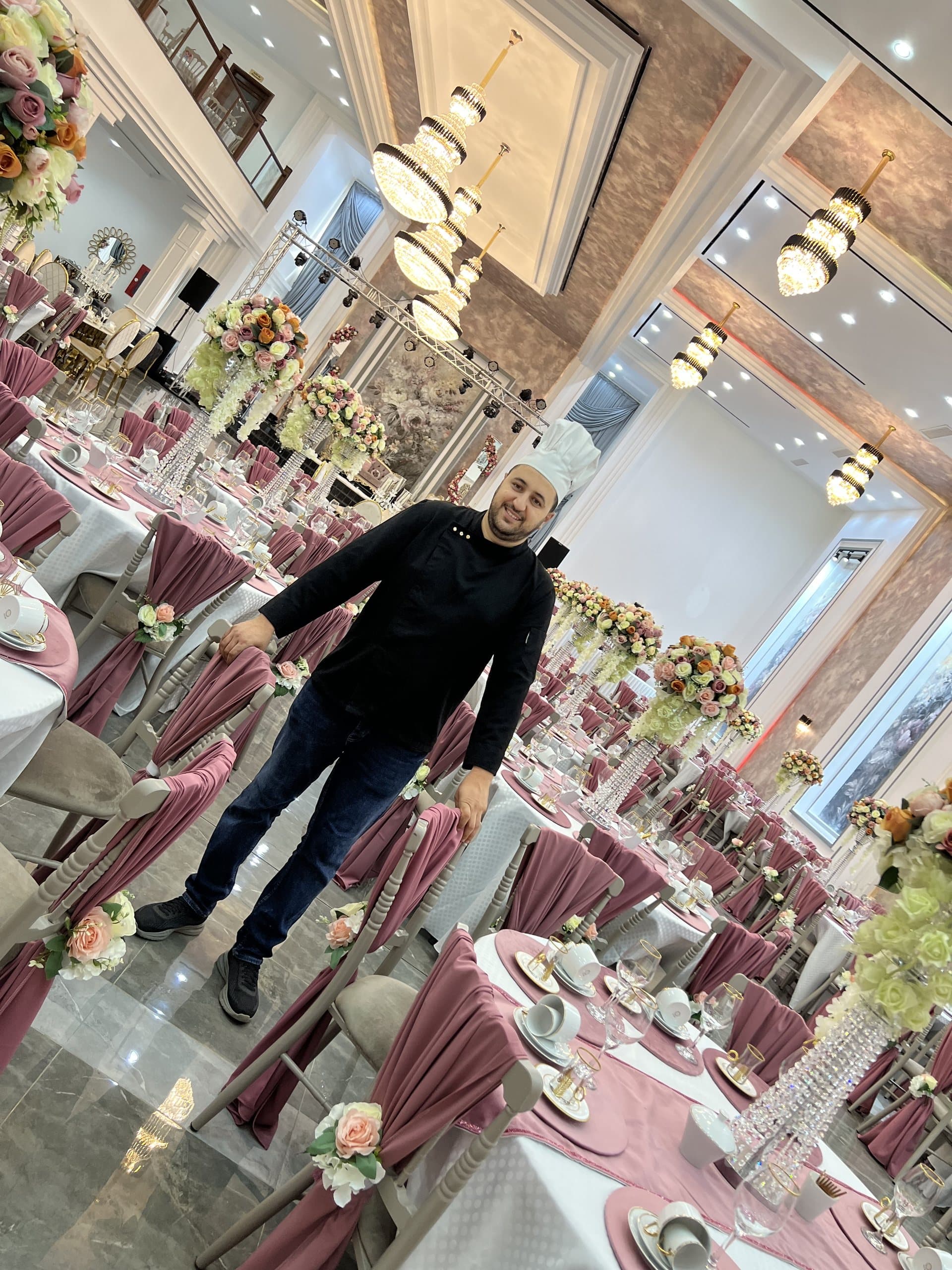 Un chef pose dans une salle de mariage décorée de fleurs et de nappes roses.