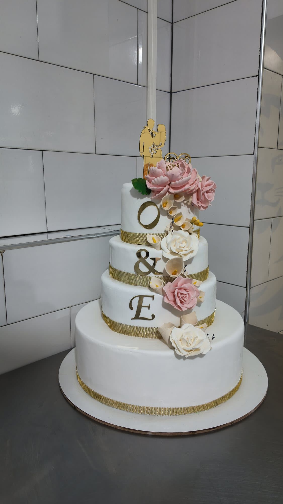 Une pièce montée de mariage à quatre étages, blanche et dorée, ornée de fleurs en sucre roses et blanches et des initiales O & E.