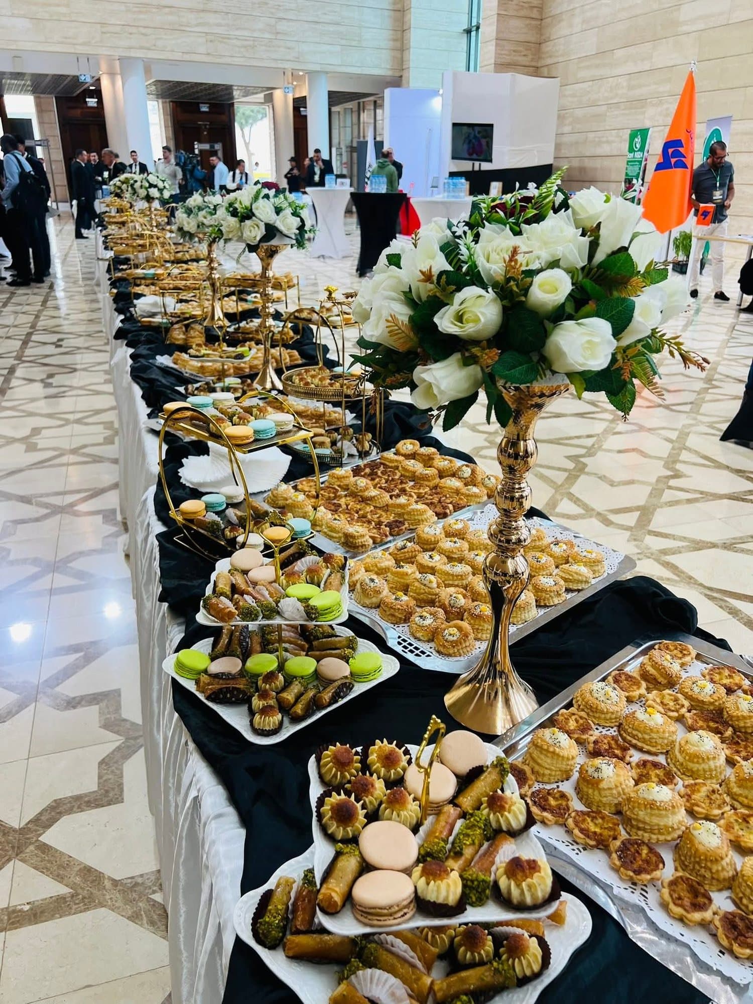 Un long buffet de pâtisseries, macarons et bouquets de roses blanches dans un grand hall.