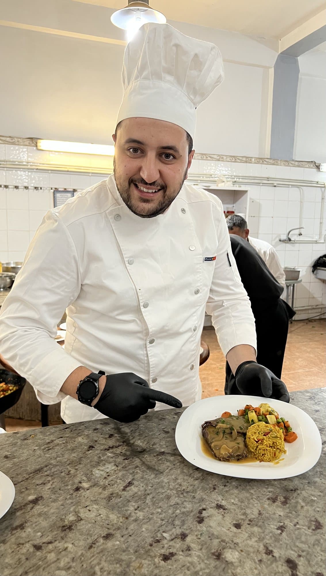 Un chef de Dar Lahbab, souriant, présente une assiette de viande en sauce, riz et légumes.