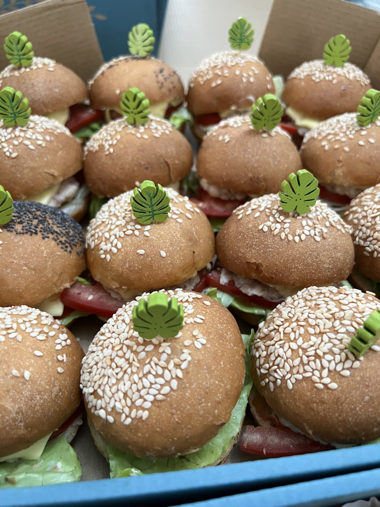 Des mini-burgers au sésame, piqués d'une feuille décorative, rangés en boîte.