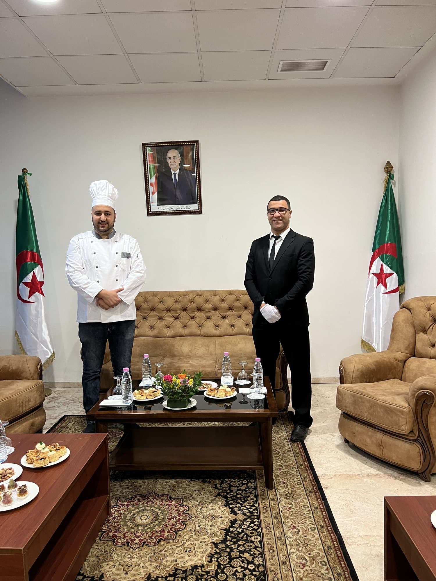 Un chef et un maître d'hôtel en gants blancs dans un salon, devant une table de douceurs.